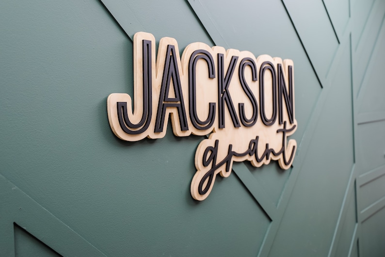 May include: A custom wall sign featuring the name JACKSON in bold, black, sans-serif capital letters layered over a light wood backing, with the name grant in a black, cursive script font layered below, creating a three-dimensional effect.