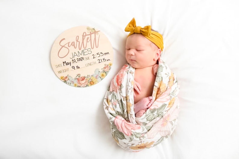 May include: A newborn baby wrapped in a floral swaddle, wearing a mustard yellow headband. A wooden sign with the name "Scarlett James" and birth details is next to the baby. The sign includes the date, time, weight (9 lbs), and length (21.5 inches).