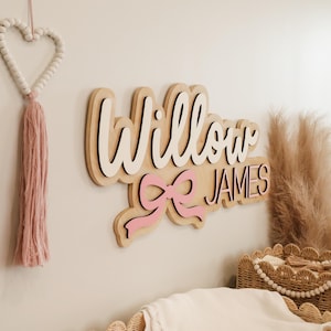May include: A layered wooden wall sign featuring the name Willow in white script lettering above the name JAMES in black block letters, accented by a pink bow graphic. The sign is mounted on a light-colored wall with a pink beaded heart tassel.