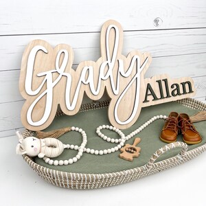 May include: Wooden name sign with the name "Grady" in a layered design, and the name "Allan" in black. The sign is displayed with baby shoes, a beaded garland, and a toy in a woven basket with a green liner.