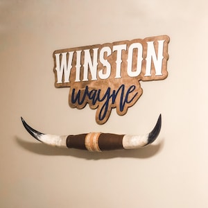 May include: A wooden wall sign featuring the name WINSTON in white block letters and the name wayne in dark blue script, positioned above a pair of long, curved animal horns wrapped in brown leather and light-colored textured material at the center.
