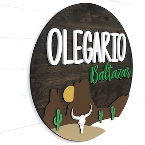 K&ouml;nnte beinhalten: Ein rundes Holzschild mit einer W&uuml;stenlandschaft mit einem Kaktus, einer Bergkette, einer Sonne und einem Bullensch&auml;del. Der Name "OLEGARIO Baltazar" ist in wei&szlig;en 3D-Buchstaben auf dem Schild geschrieben.