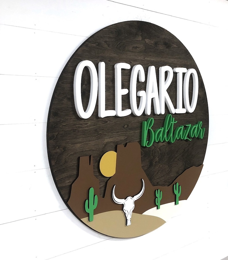 K&ouml;nnte beinhalten: Ein rundes Holzschild mit einer W&uuml;stenlandschaft mit einem Kaktus, einer Bergkette, einer Sonne und einem Bullensch&auml;del. Der Name "OLEGARIO Baltazar" ist in wei&szlig;en 3D-Buchstaben auf dem Schild geschrieben.
