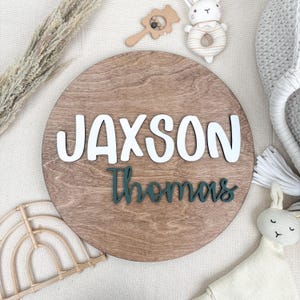 May include: A round wooden sign with the name "Jaxson" in white letters and the name "Thomas" in green script. The sign is surrounded by baby toys, including a rattle, a stuffed bunny, and a rainbow-shaped decoration.