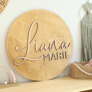 May include: A circular wooden sign featuring the name "Liana Marie". The name "Liana" is in a soft pink hue, while "MARIE" is in white. The sign is crafted from light-toned wood, suitable for a nursery.