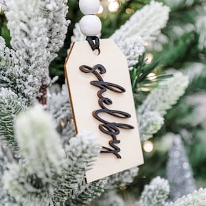 May include: A white wooden ornament with the word "shelter" written in black calligraphy on it. The ornament is hanging on a snowy artificial Christmas tree.