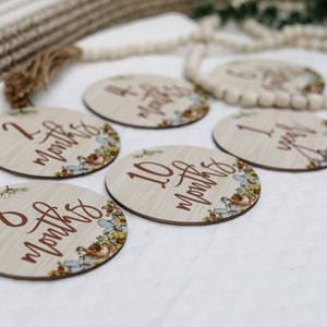 May include: Wooden milestone discs with floral accents and months/year inscriptions. The discs are light-colored with brown text and floral designs in shades of blue, yellow, and brown. A decorative wooden bead garland is in the background.
