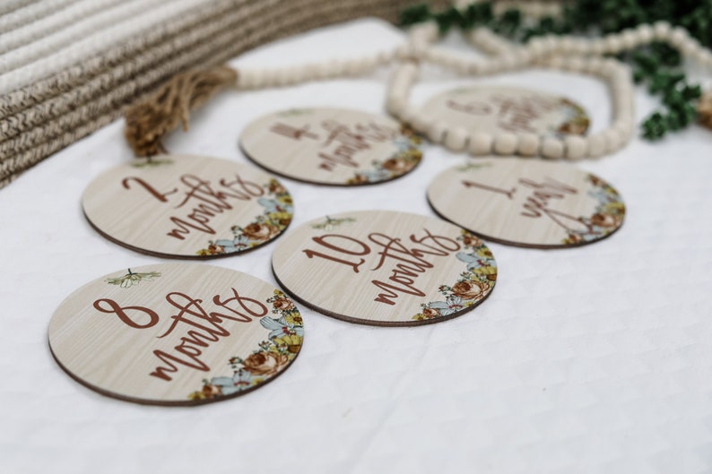 May include: Wooden milestone discs with floral accents and months/year inscriptions. The discs are light-colored with brown text and floral designs in shades of blue, yellow, and brown. A decorative wooden bead garland is in the background.
