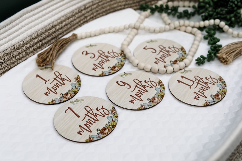 May include: A set of six wooden monthly milestone discs. Each disc features a floral design and the month number in a dark brown script. The discs are arranged on a white surface with a decorative beaded garland.