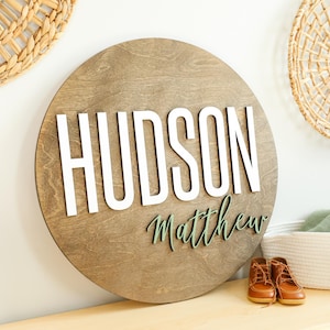 May include: A round wooden sign with the name "HUDSON" in large white letters and the name "Matthew" in green script. The sign is brown and sits on a wooden surface with brown leather baby shoes.