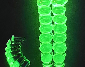 May include: Bright green, glowing ear gauges in a vertical arrangement. The image shows two columns of circular gauges and a curved row of gauges, all with a translucent appearance against a dark background. The gauges appear to be made of glass or a similar material.
