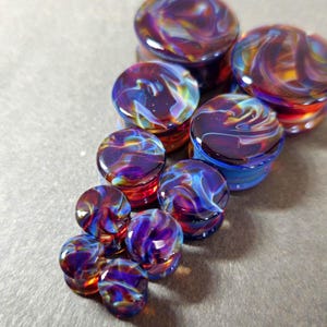 May include: A collection of circular glass ear plugs in varying sizes, featuring a swirling pattern of deep purple, vibrant blue, and fiery orange hues. Each piece has a smooth, polished surface with a glossy finish, reflecting light across the colours.