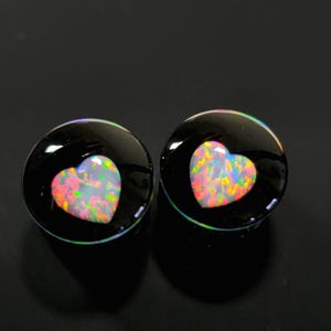 Puede incluir: Un par de tapones redondos negros con un corazón de ópalo. Los ópalos muestran una vibrante gama de colores, incluyendo rosa, amarillo y verde. Los tapones tienen un acabado brillante y están sobre un fondo oscuro.