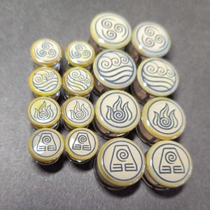 May include: A collection of ear gauges with various symbols representing air, water, fire, and earth. The gauges have a cream-colored face with black line art and a silver-colored metal base. The symbols are circular and stylized.