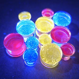 Puede incluir: Una colección de coloridos y brillantes dilatadores para orejas en tonos azul, amarillo y rosa. Los tapones tienen un diseño claro y redondeado con una textura interna visible, sobre un fondo oscuro.