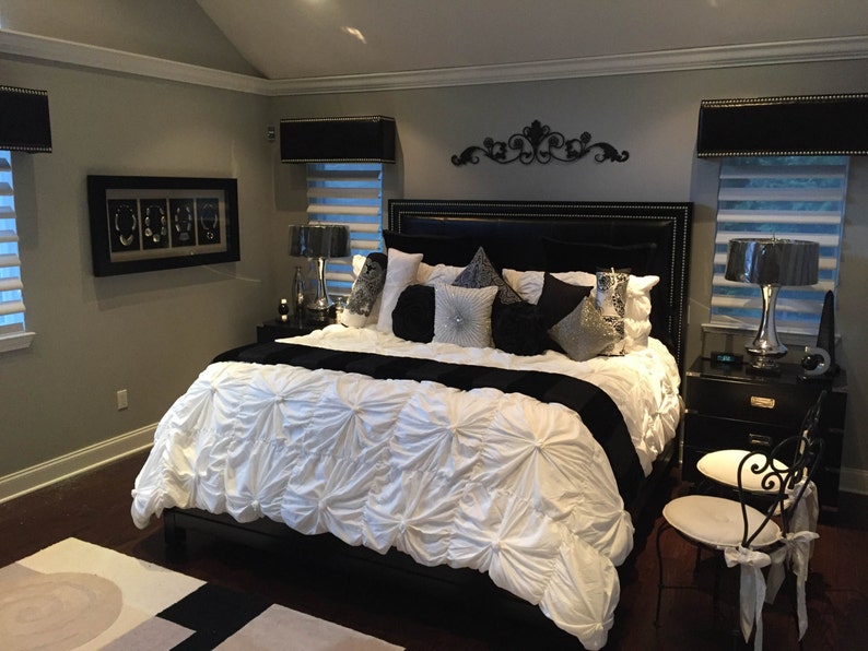 May include: A black and white bedroom with a large bed with a white comforter and black pillows. There are two black lamps on nightstands next to the bed. The walls are painted a light gray color.