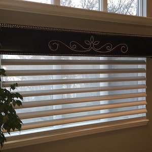 May include: A white window blind with a brown valance featuring a decorative design with silver accents. The valance is upholstered with a dark brown fabric and has a row of silver studs along the bottom edge.