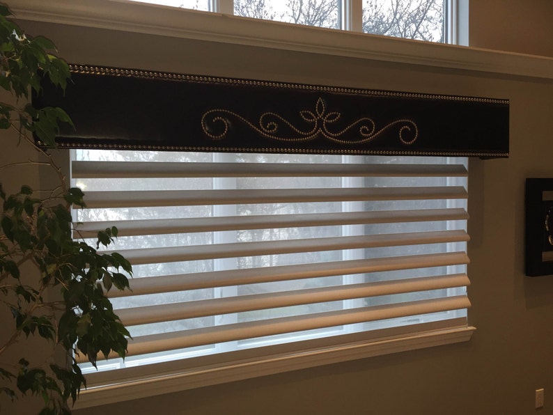 May include: A white window blind with a brown valance featuring a decorative design with silver accents. The valance is upholstered with a dark brown fabric and has a row of silver studs along the bottom edge.