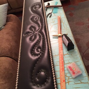 May include: A black leather headboard with a decorative stitched design and silver bead trim. The headboard is being worked on with tools and a ruler.