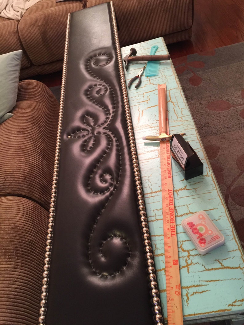May include: A black leather headboard with a decorative stitched design and silver bead trim. The headboard is being worked on with tools and a ruler.