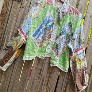 May include: A long-sleeve button-down shirt featuring a vibrant, multi-coloured collage print of Australian landscapes, landmarks, and flora. The shirt has a light green base colour, a standard collar, and three visible round buttons down the front.