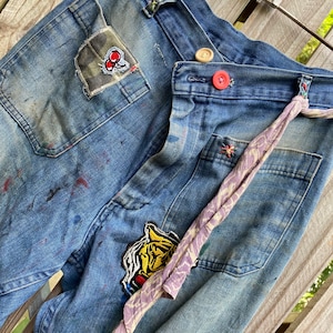 May include: Blue denim jeans with multiple patches, including a tiger, a plaid design, and a red cherry. The jeans have a faded look and are decorated with a purple and white patterned fabric tie.