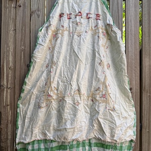May include: A white apron with the word "PEACE" embroidered in red letters. The apron has floral embroidery and a green and white checked bottom hem. It hangs on a wooden hanger against a wooden fence.