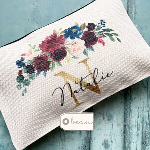 Könnte beinhalten: Eine weiße Canvas-Kosmetiktasche mit einem Blumendesign in den Farben Burgunderrot, Blau und Grün. Die Tasche hat einen goldenen Buchstaben "N" und den Namen "Natalie" in schwarzer Kursivschrift.