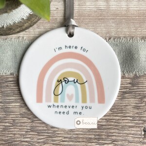 May include: A white ceramic ornament with a rainbow design and the text "I'm here for you whenever you need me." The ornament has a small heart in the center of the rainbow and a white ribbon for hanging.