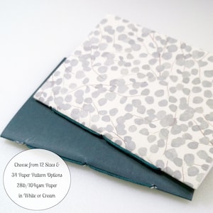 May include: A notebook with a white cover featuring a gray leaf pattern layered over a dark teal cover. The text on the image reads: Choose from 12 Sizes & 34 Paper Pattern Options 28lb/104gsm Paper in White or Cream. The binding is visible.