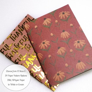 May include: Two notebooks with rectangular covers. One cover features a repeating pattern of orange flowers on a dark red background. The other cover has the gold foil text THANKFUL repeated vertically and includes a circular white label.