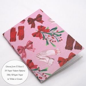 May include: A notebook with a pink cover featuring a repeating pattern of red bows, red gloves, white gloves, and green holly sprigs with red berries. A circular white label includes the text: Choose from 12 Sizes & 34 Paper Pattern Options 28lb/104gsm Paper in White or Cream.