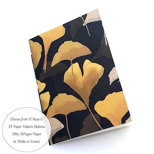 May include: A small notebook with a black cover featuring a pattern of gold ginkgo leaves. The text on the cover reads "Choose from 12 Sizes & 24 Paper Pattern Options 28lb/104gsm Paper in White or Cream".