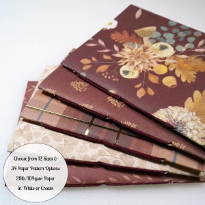 May include: A stack of four folded paper booklets with stitched spines. The top cover features a dark burgundy background with floral and mushroom illustrations. Below it, a plaid pattern with gold foil lines, followed by a light beige leaf print.