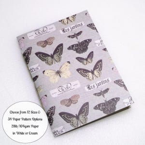 May include: A notebook with a light gray cover featuring a repeating pattern of various butterflies and vintage-style labels with the text "Les jardins" and "Chose". The cover includes text stating: Choose from 12 Sizes & 34 Paper Pattern Options 28lb/104gsm Paper in White or Cream.