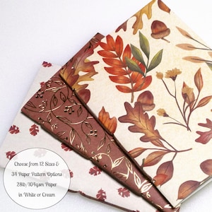 May include: Three stacked paper sheets featuring autumn-themed designs. One sheet has a cream background with orange and brown leaves and acorns. Another is a solid brown with gold foil vine patterns. The third is white with small red leaf prints. Text reads: Choose from 12 Sizes & 34 Paper Pattern Options 28lb/104gsm Paper in White or Cream.