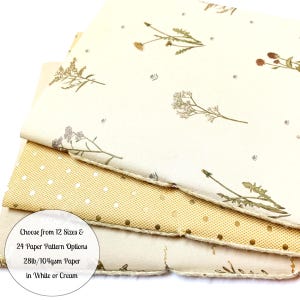 May include: Stack of four sheets of paper with different patterns. The top sheet has a floral pattern with dried flowers on a cream background. The second sheet has a gold polka dot pattern on a cream background. The third sheet has a gold polka dot pattern on a cream background. The bottom sheet has a floral pattern with dried flowers on a cream background. The text on the bottom sheet reads: "Choose from 12 Sizes & 24 Paper Pattern Options 28lb/104gsm Paper in White or Cream".