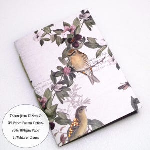 May include: A notebook with a white cover featuring a botanical print of green leaves, purple flowers, and two birds. The cover includes faint text and a circular white label that reads: Choose from 12 Sizes & 34 Paper Pattern Options 28lb/104gsm Paper in White or Cream.