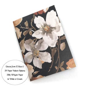 May include: A rectangular notebook featuring a black background with a watercolor floral pattern in shades of white, tan, and brown. Text on the cover reads: Choose from 12 Sizes & 34 Paper Pattern Options 28lb/104gsm Paper in White or Cream.