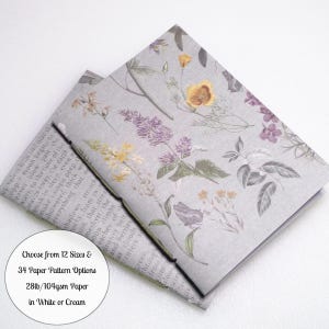 May include: A rectangular notebook with a light gray cover featuring a botanical print of yellow and purple flowers, green leaves, and small insects. A portion of the notebook shows text printed on the paper. A circular white label displays the text: Choose from 12 Sizes & 34 Paper Pattern Options 28lb/104gsm Paper in White or Cream.