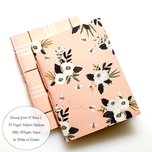 May include: Two notebooks with floral patterns and gold accents. The top notebook has a pink and white plaid pattern. The bottom notebook has a pink background with white flowers and bees. The text on the notebooks reads: "Choose from 12 Sizes & 24 Paper Pattern Options 28lb/104gsm Paper in White or Cream"