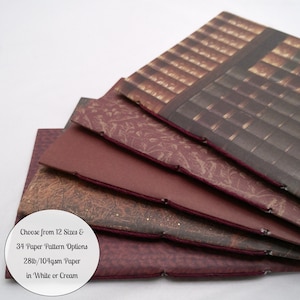 May include: A stack of five hand-bound booklets with dark red, brown, and patterned covers, featuring exposed maroon stitching along the spines. Text on the image reads: Choose from 12 Sizes & 34 Paper Pattern Options 28lb/104gsm Paper in White or Cream.