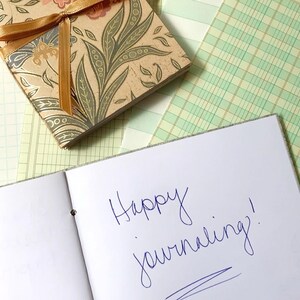 May include: An open journal with the words "Happy journaling!" written in blue ink. A decorative floral patterned box with a gold ribbon sits above the journal. The background includes green and white patterned paper.