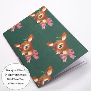 May include: A dark green notebook featuring a repeating pattern of brown fawns with pink bows and holly leaves. A circular white label includes the text: Choose from 12 Sizes & 34 Paper Pattern Options 28lb/104gsm Paper in White or Cream.