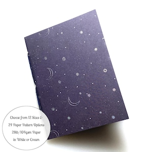May include: A dark purple notebook with a silver foil moon and star pattern. The notebook is closed and the spine is visible. The text on the cover reads "Choose from 12 Sizes & 24 Paper Pattern Options 28lb/104gsm Paper in White or Cream".