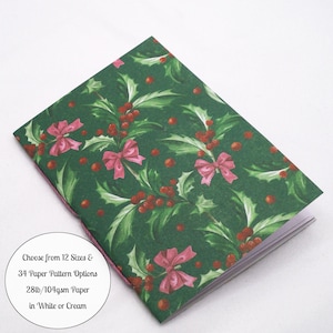 May include: A notebook with a dark green cover featuring a repeating pattern of red holly berries and pink bows. A circular white label on the cover reads: Choose from 12 Sizes & 34 Paper Pattern Options 28lb/104gsm Paper in White or Cream.