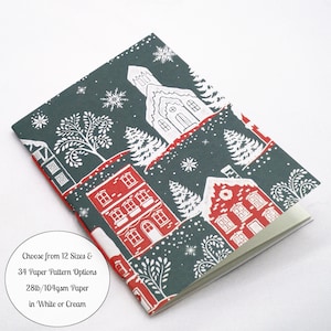May include: A notebook with a dark green cover featuring a white and red winter village print, including houses, a church, trees, and snowflakes. A circular white label states: Choose from 12 Sizes & 34 Paper Pattern Options 28lb/104gsm Paper in White or Cream.