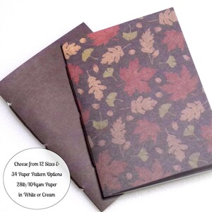 May include: Two notebooks with a dark brown cover featuring a repeating pattern of red, orange, and green autumn leaves and acorns. A circular white label includes the text: Choose from 12 Sizes & 34 Paper Pattern Options 28lb/104gsm Paper in White or Cream.