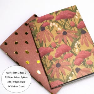 May include: Two notebooks with rectangular covers. One cover features a floral pattern with red flowers and green stems on a tan background. The other cover is dark brown with a repeating pattern of small, shiny gold foil acorn shapes.