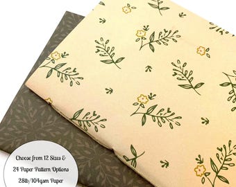 May include: Two notebooks, one with a cream-colored floral pattern and the other with a dark green leaf pattern. The cream notebook has a design of green leaves and yellow flowers. The text on the image reads: Choose from 12 Sizes & 24 Paper Pattern Options 28lb/104gsm Paper in White or Cream.